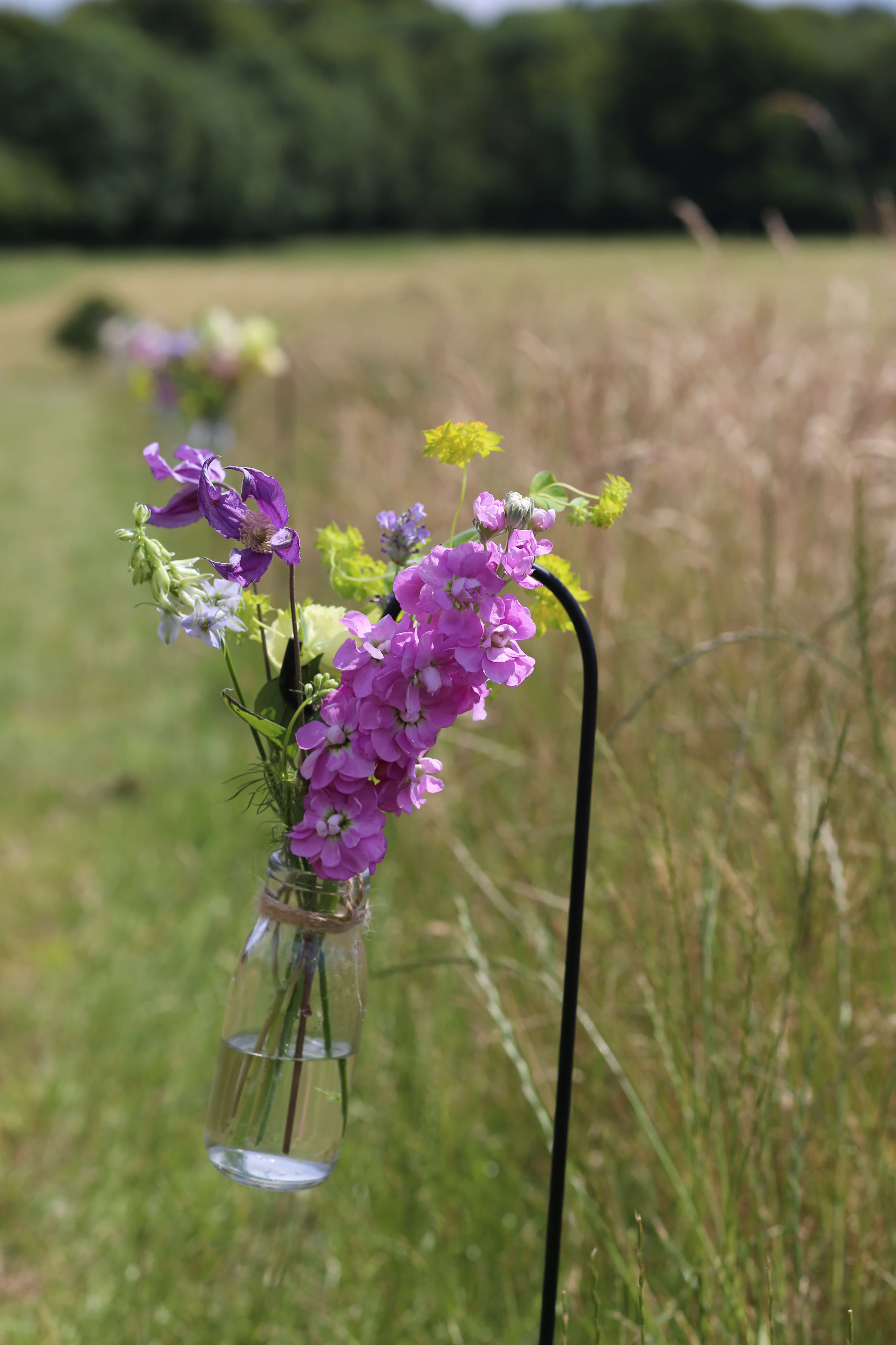 meadow style flowers in a glass bottle hanging from a shepherd hook
