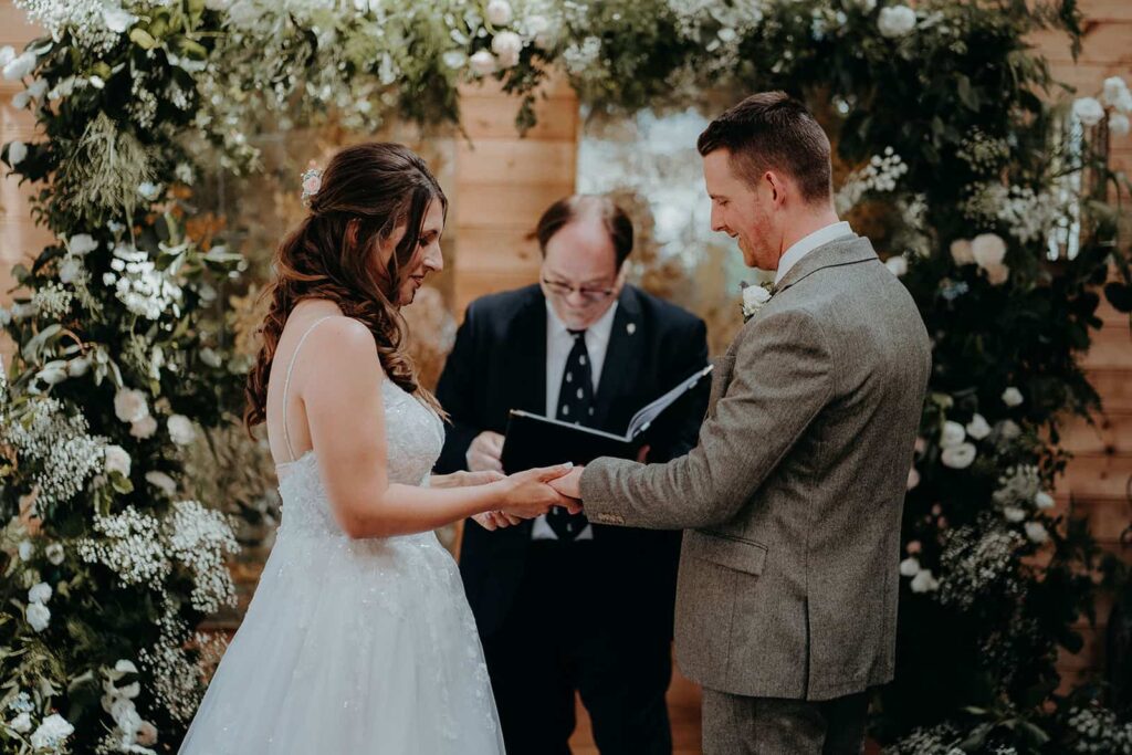 bride and groom standing taking their vows underneath a floral arch in the ceremony barn at chafford park estate